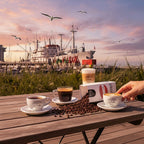 Weiße Kaffeetüte des Cuxhavener Kaffeeroesters mit der Aufschrift Espresso Alte Liebe auf Terrassentisch und vier Kaffeespezialitäten vor Hafenkulisse an der Nordsee 

