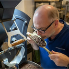 Eigentümer an Kaffeeröstmaschine, der die Qualität von frisch geröstetem Kaffee sensorisch prüft