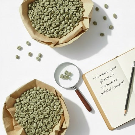 Zwei Schalen mit besten Rohkaffeebohnen sowie einer Lupe zur Qualitätsprüfung und einem Notizbuch mit der Eintragung: schonend und gänzlich chemiefrei entkoffeiniert!
