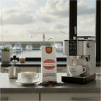 Weiße Kaffeetüte des Cuxhavener Kaffeeroesters mit der Aufschrift Crema Neue Liebe mit Kaffeevollautomat, Cappuccino und Fenster mit Nordseeblick
