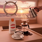 Weiße Kaffeetüte des Cuxhavener Kaffeeroesters mit der Aufschrift Kugelbake Blend  mit Filterkaffekanne in Schiffsküche und Fenster mit Nordseeblick bei Sonnenuntergang.
