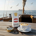 Weiße Kaffeetüte des Cuxhavener Kaffeeroesters mit der Aufschrift Lotsenmischung  auf Tisch mit Cappuccino auf einem Segelschiff und  Nordseeblick
