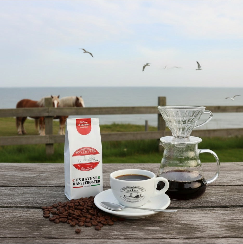 Weiße Kaffeetüte des Cuxhavener Kaffeeroesters mit der Aufschrift Papua Neuguinea auf Gartentisch mit Blick auf Pferdekoppel und Nordsee