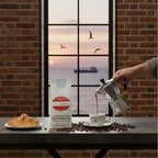 Weiße Kaffeetüte des Cuxhavener Kaffeeroesters mit der Aufschrift Peru mit Bialetti und frisch gebrühtem Espresso  und Fenster mit Nordseeblick
